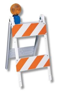 Portable barricade with orange-and-white diagonal panels and a yellow warning beacon on top, used to block access at a worksite or construction zone.